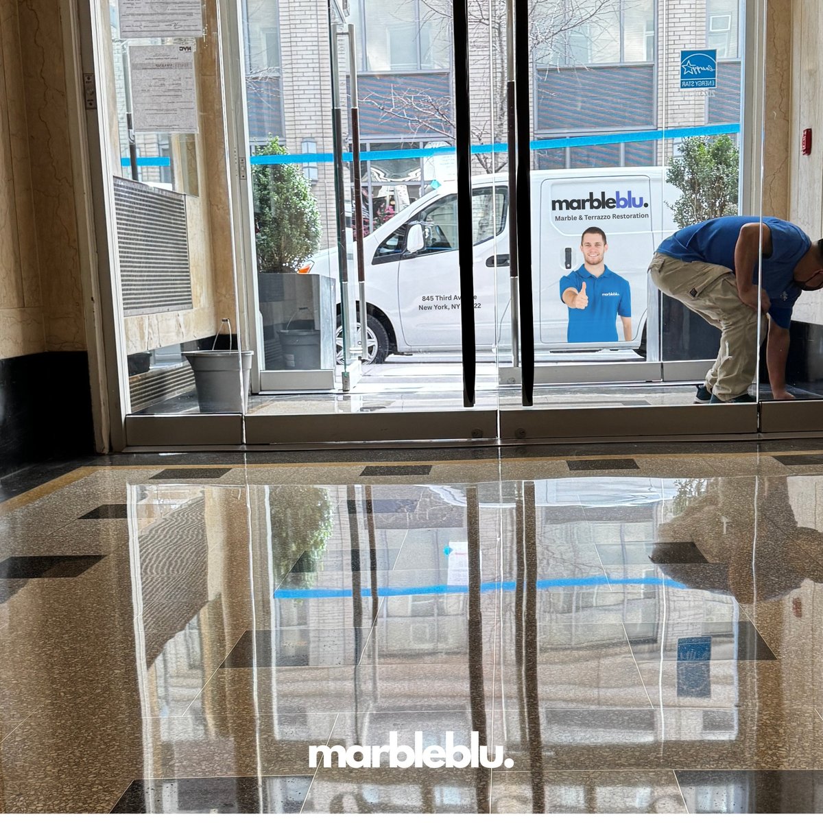 Interior view of Marbleblu storefront with glass doors, polished floor reflecting storefront signage, and white delivery van visible outside