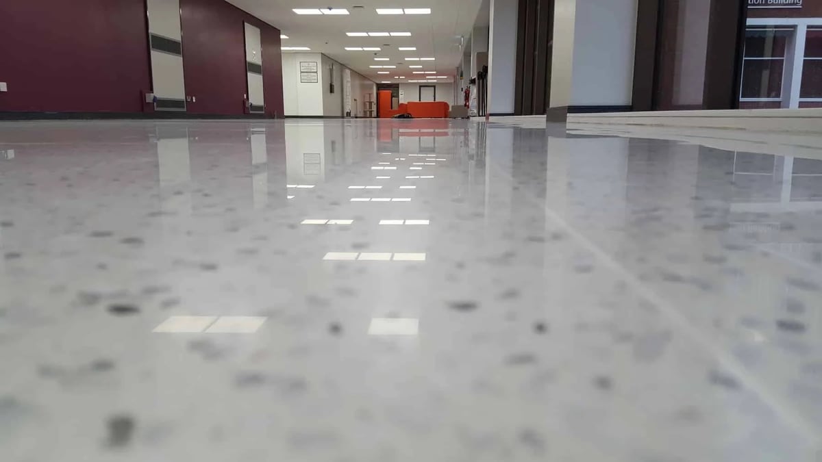 Empty modern office corridor with polished concrete floor, ceiling lights, burgundy and white walls, and red chairs visible in background