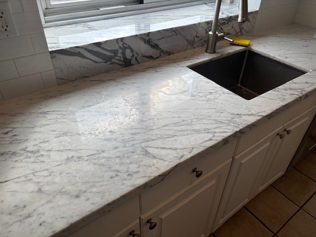 Marble kitchen countertop with black square sink and chrome faucet, white cabinetry below, bright window behind — before restoration